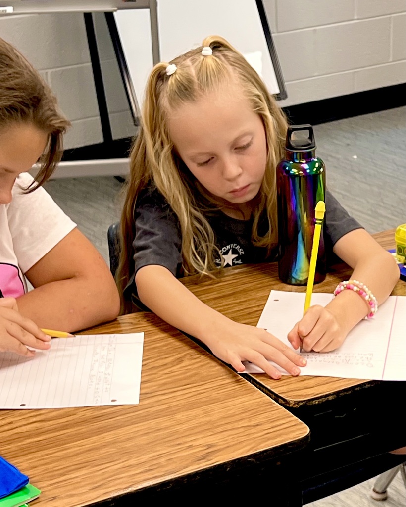 Two students sit at desks, writing. The girl in front wears a bracelet and uses a yellow pencil.