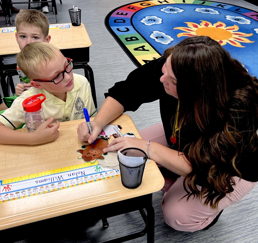 A teacher kneels next to a child, assisting with an activity at a school desk.