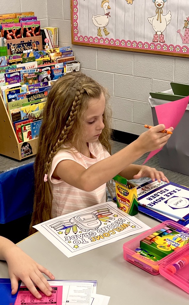 A girl sits at a table, holding a pencil and working on a coloring page. Various items surround her.