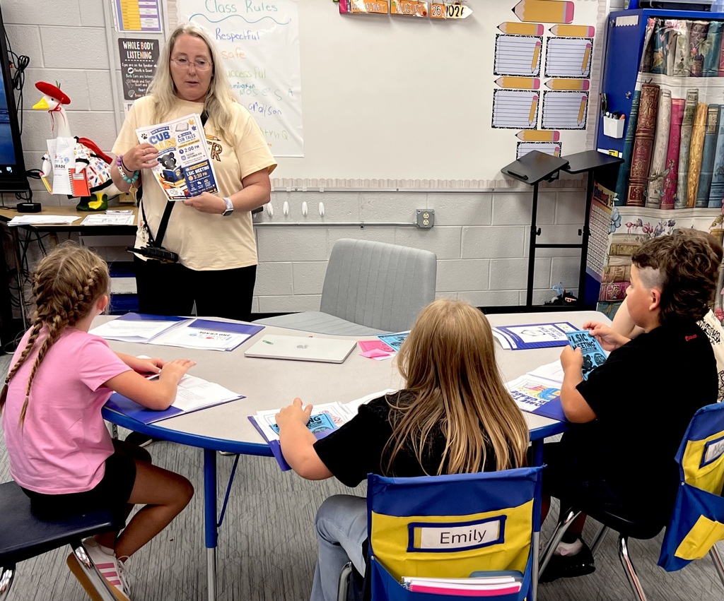 A teacher, standing, holds a book and addresses students seated around a table. They have papers and books.