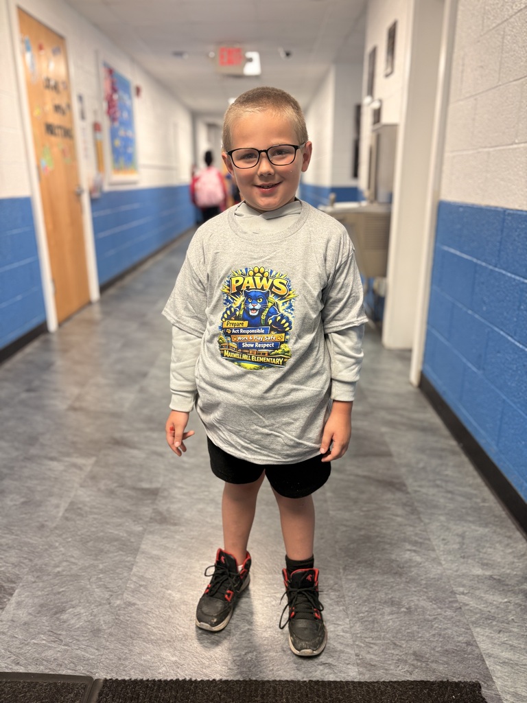 A young boy in a long-sleeve shirt with a cartoon character, glasses, and sneakers stands in a school hallway.