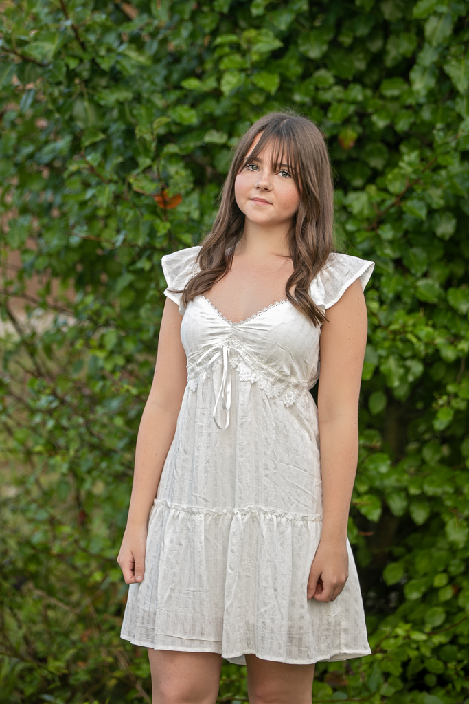 A person in a white dress with ruffled shoulders stands in front of a bush.