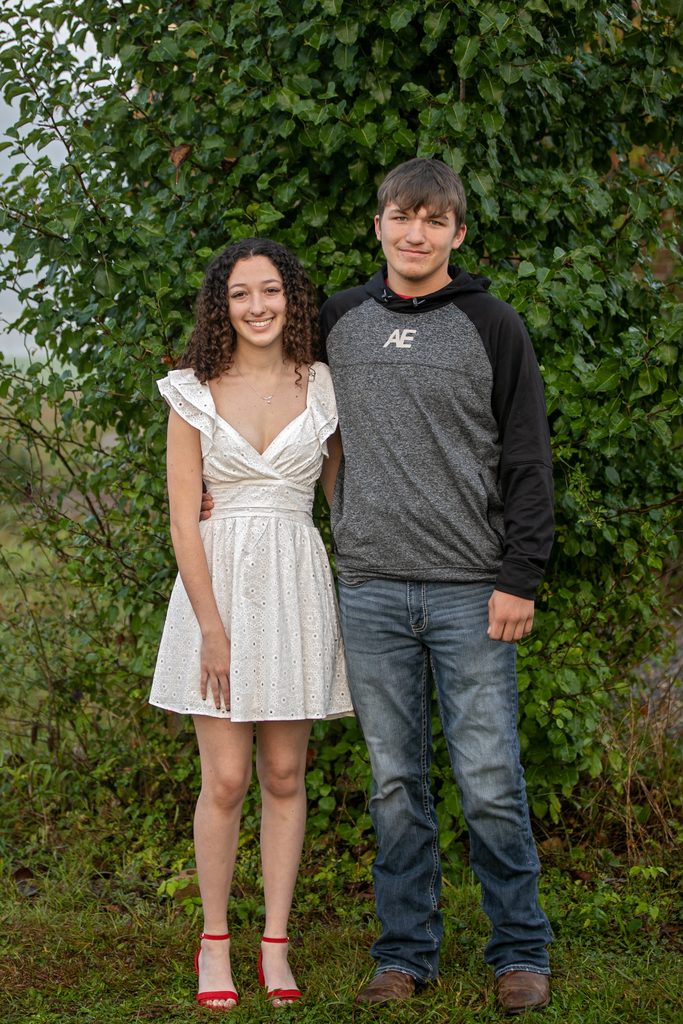 A couple in matching poses, woman in white dress, man in gray hoodie, standing in a field with a green bush background.