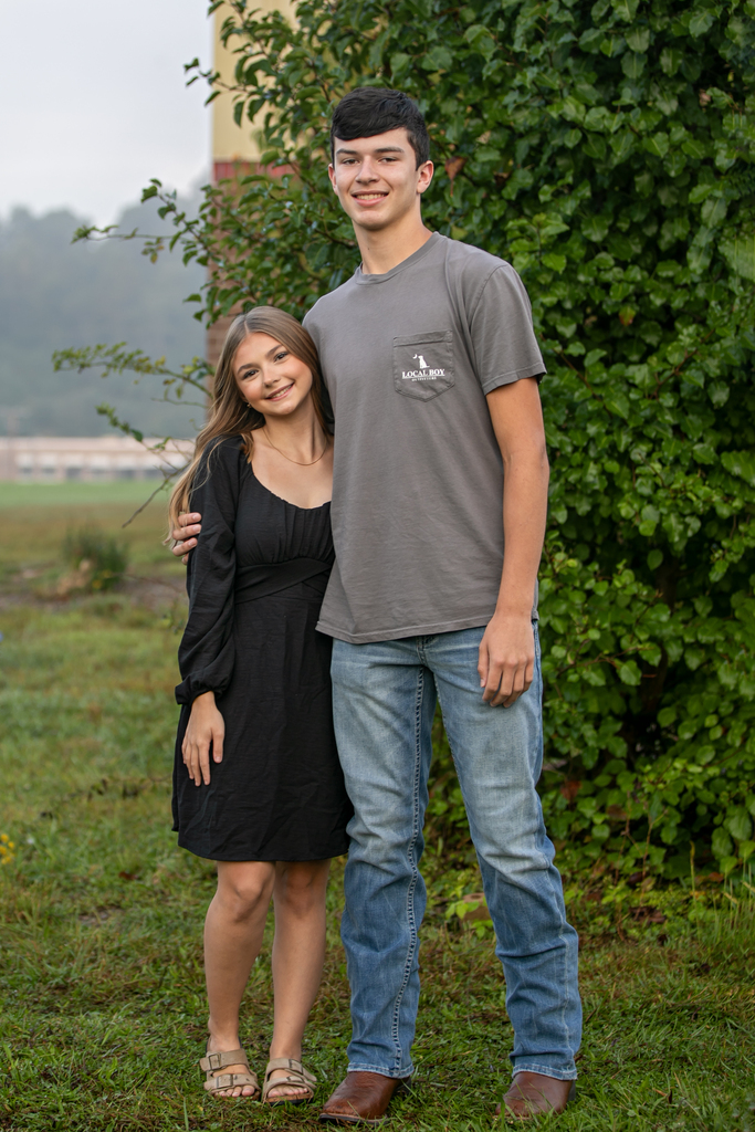 A couple stands in front of a green bush, both smiling. The man wears a gray shirt, jeans, and brown shoes. The woman wears a black dress, sandals, and a necklace.