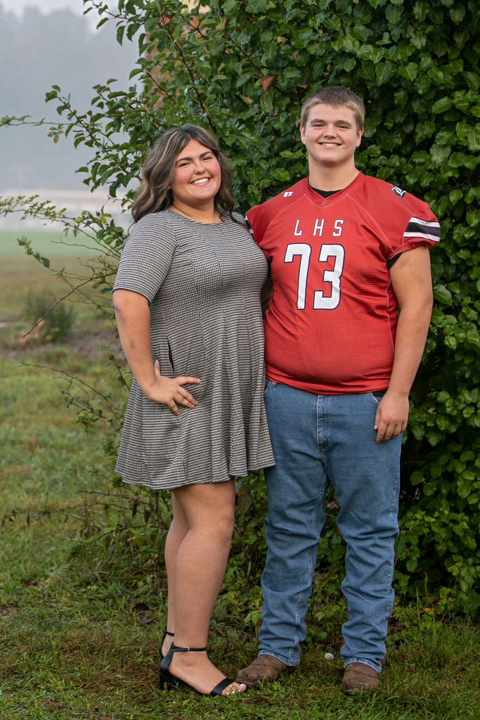 A young woman and man in a football jersey stand together, posing for a photo. They are outside with green shrubs behind them.