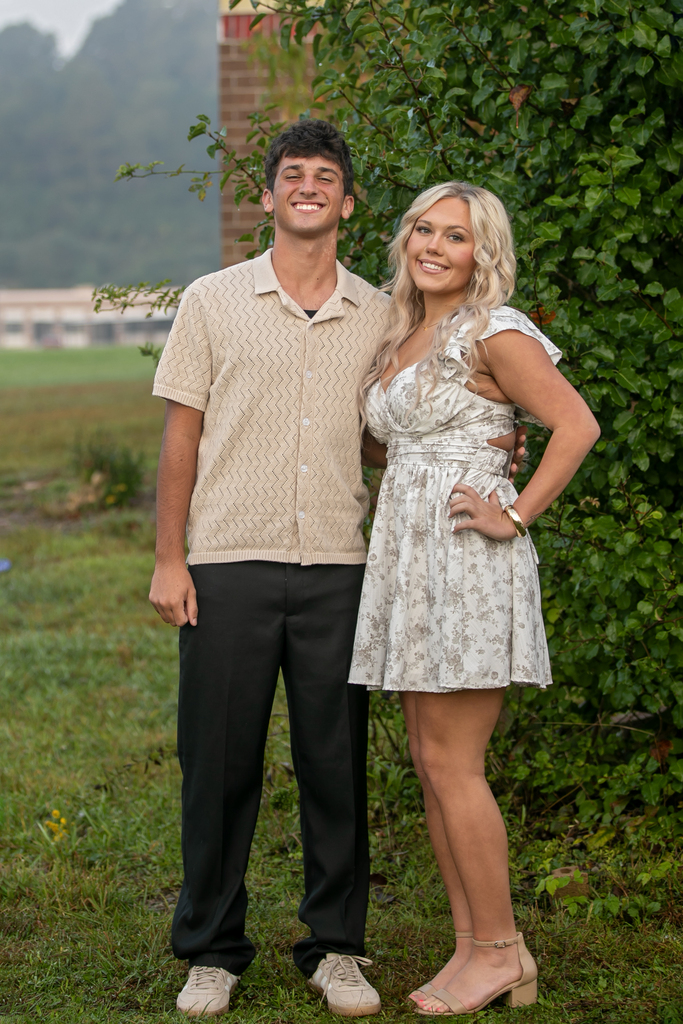 A couple stands on grass, smiling, with a hedge and distant buildings in the background.