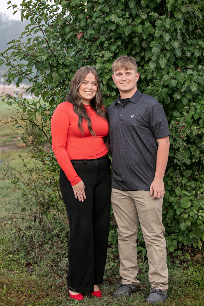 A woman in a red top and a man in a gray polo stand together in front of a green bush.