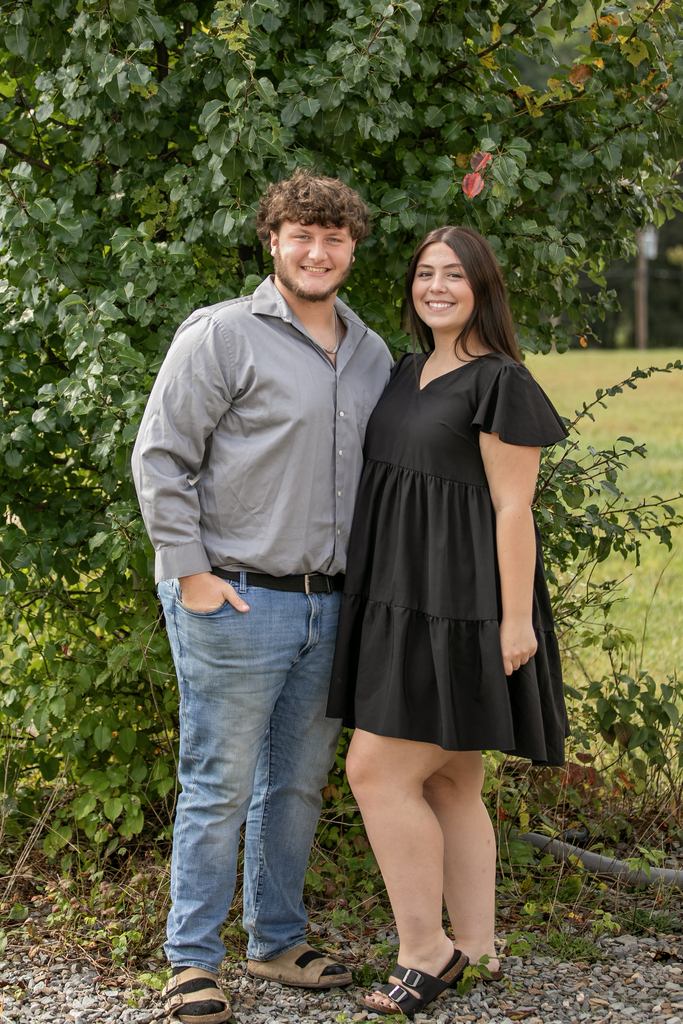 A couple stands close, smiling at the camera. He wears a gray shirt and jeans. She wears a black dress.