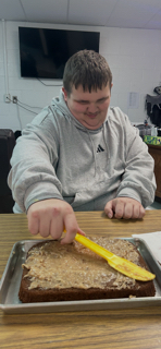 A person in a gray hoodie sitting at a table, using a yellow spatula to spread frosting on a chocolate cake.