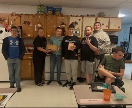 A group of people stands in a kitchen with a table. One holds a trophy. The kitchen has cabinets and appliances.