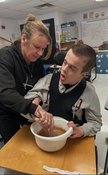A woman and a boy in a wheelchair in a classroom. The woman helps the boy mix something in a bowl.