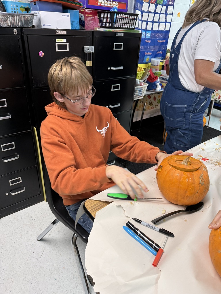 A young boy with glasses and an orange hoodie is carving a pumpkin on a table.