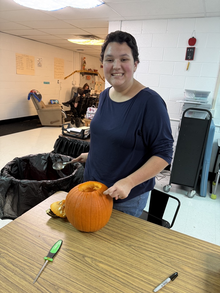 A person is smiling while holding a pumpkin with a knife in a room with chairs.