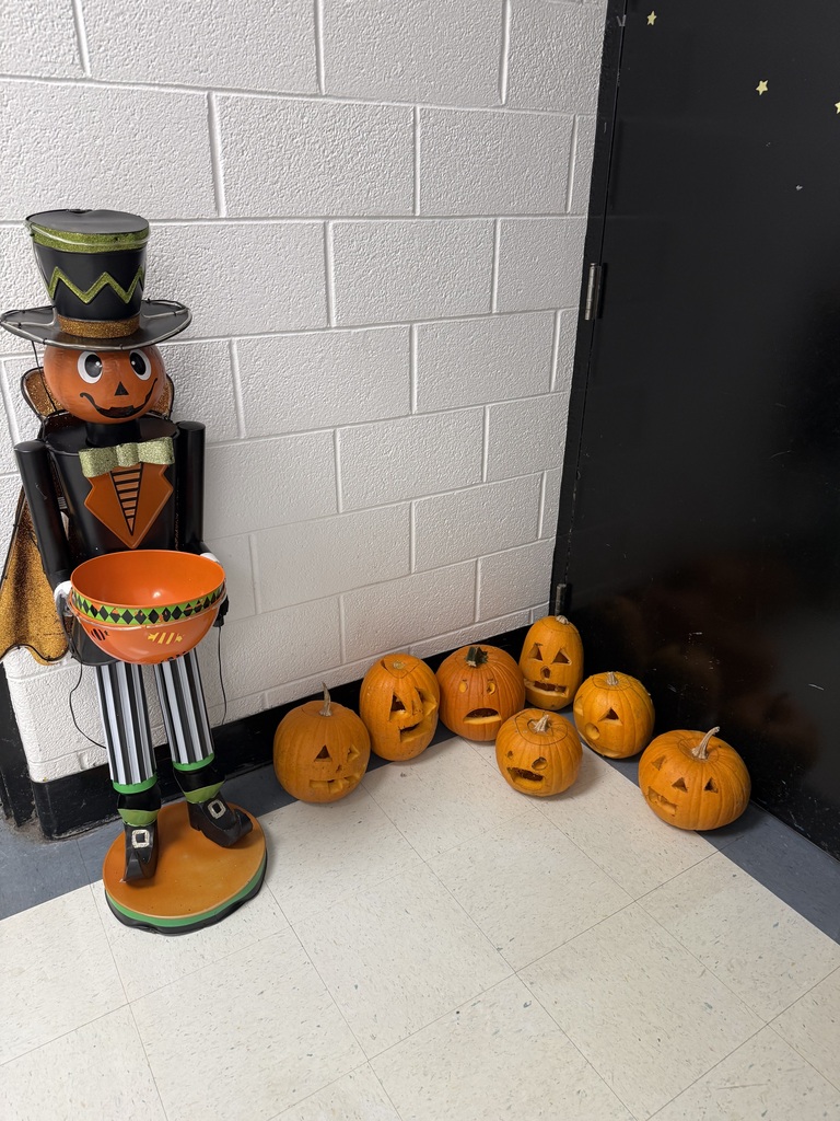 A pumpkin-themed figure stands next to a white brick wall, holding a bowl. Eight carved pumpkins are on the floor.