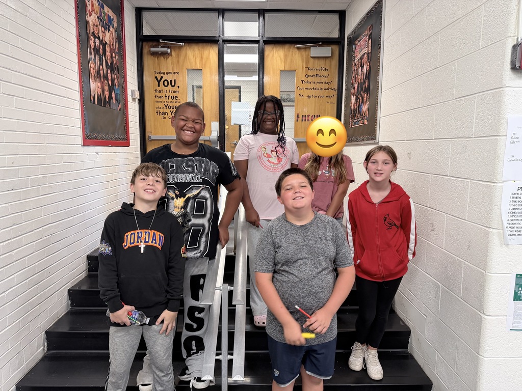 Five kids stand on stairs in a school hallway. One holds a smiley face. Behind them are doors and wall posters.