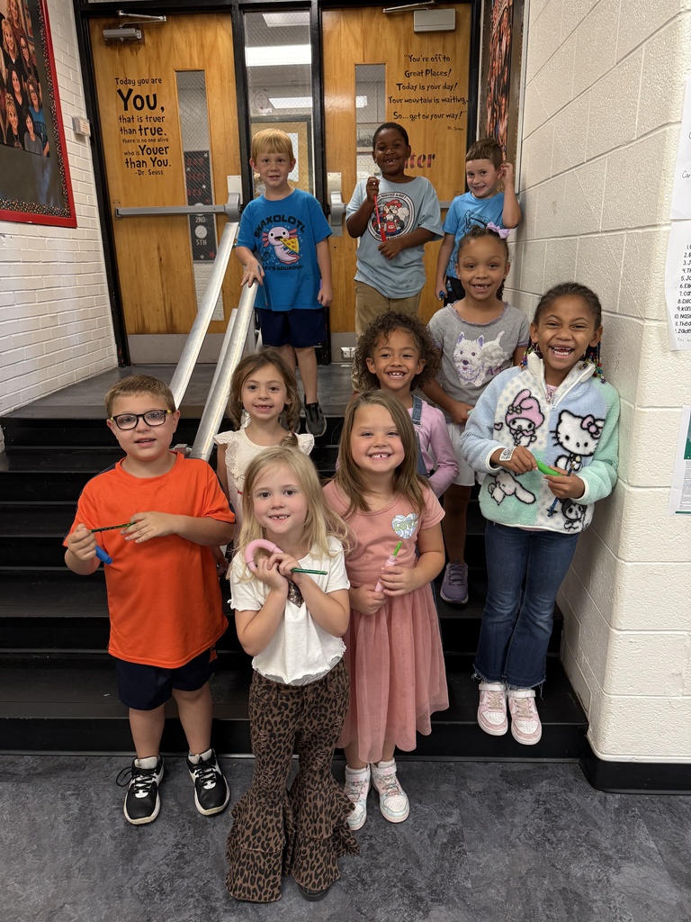 Nine children, some wearing glasses, pose for a photo in a school hallway. The background includes a white wall and wooden doors.