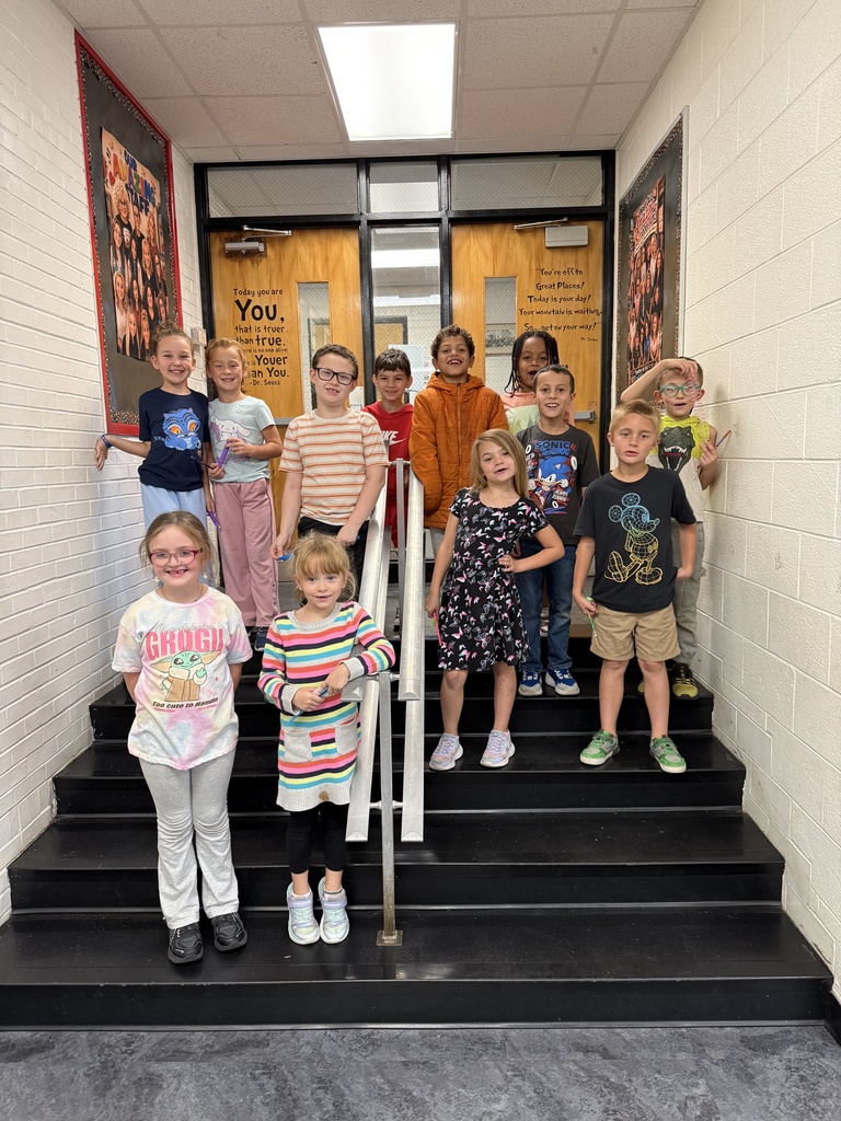 Nine children in a school hallway, standing on stairs and smiling, with a white wall and doors in the background.