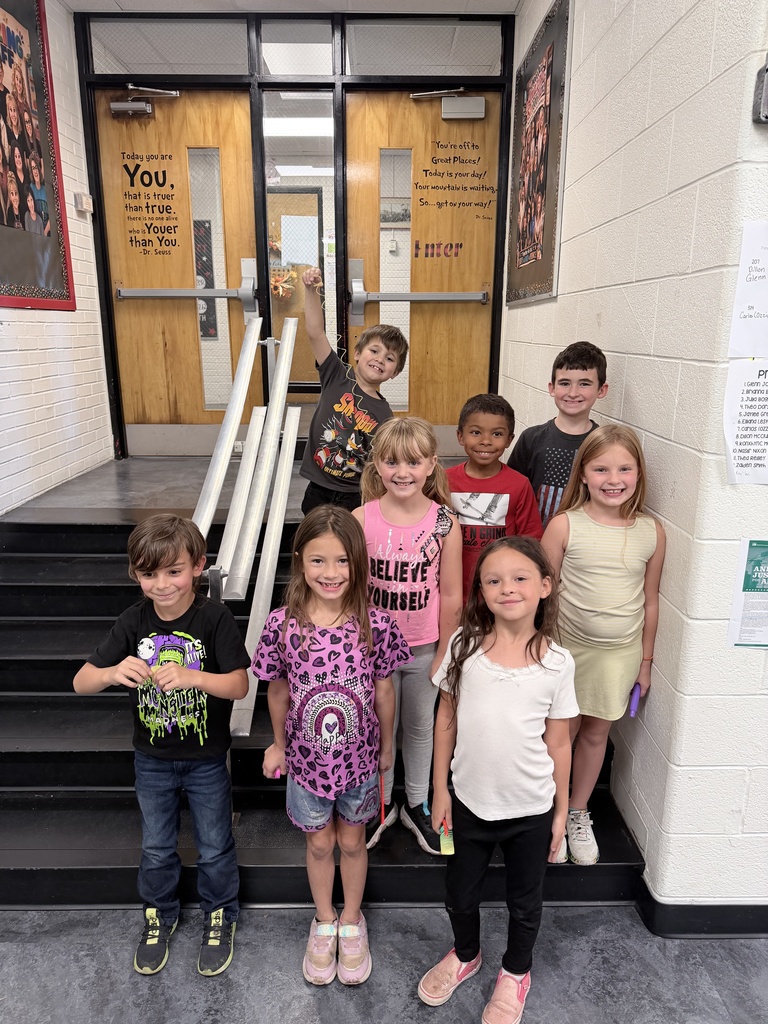 Nine children stand together on the stairs in front of a school entrance. The walls have a white background with framed posters.