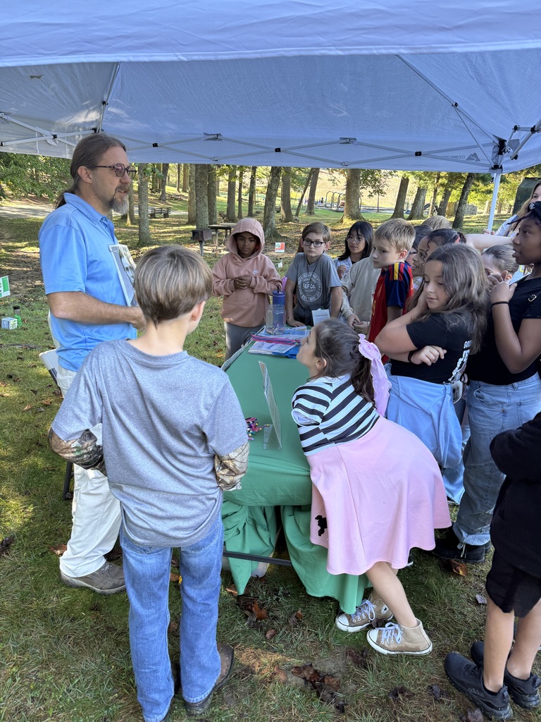 A group of children and an adult male under a tent. A green table holds items. They are outdoors with trees.
