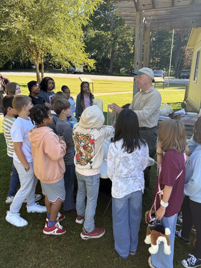 Several children gather around a man in front of a yellow building, surrounded by trees and grass.