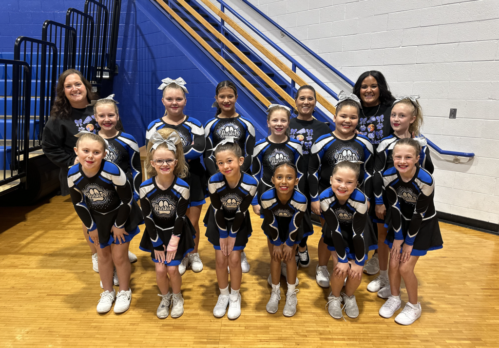 Nine girls in cheerleading uniforms and white shoes pose in a gym with two adults.