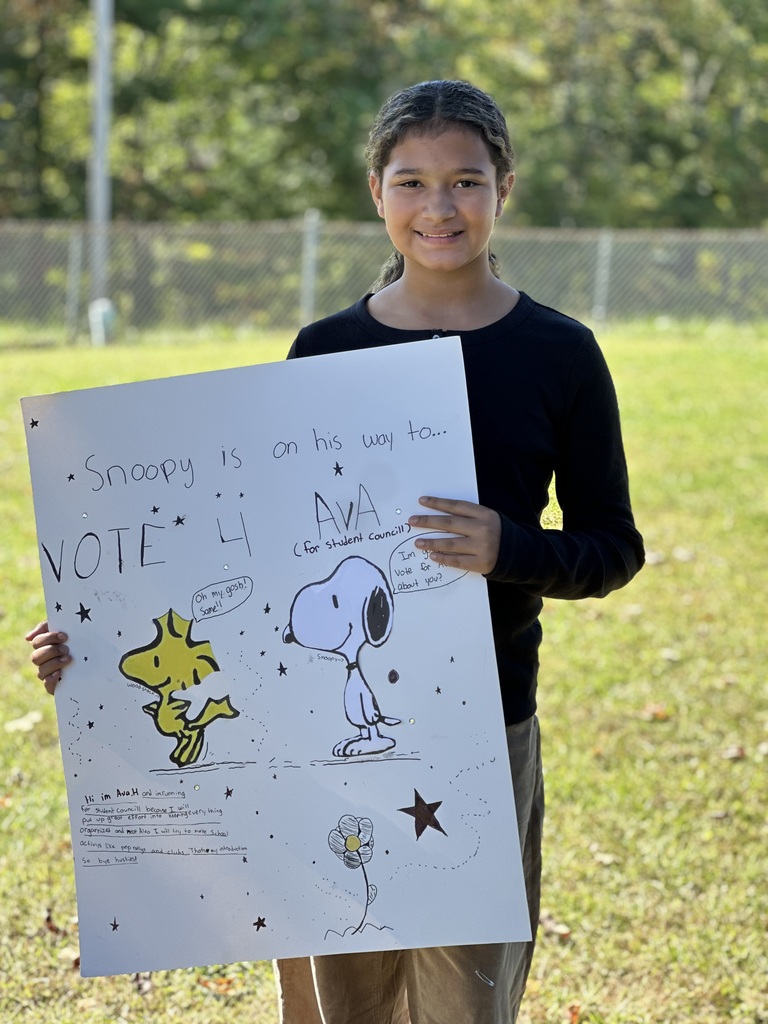Child smiling, holding a large white poster with text and Snoopy drawings, in a grassy area.