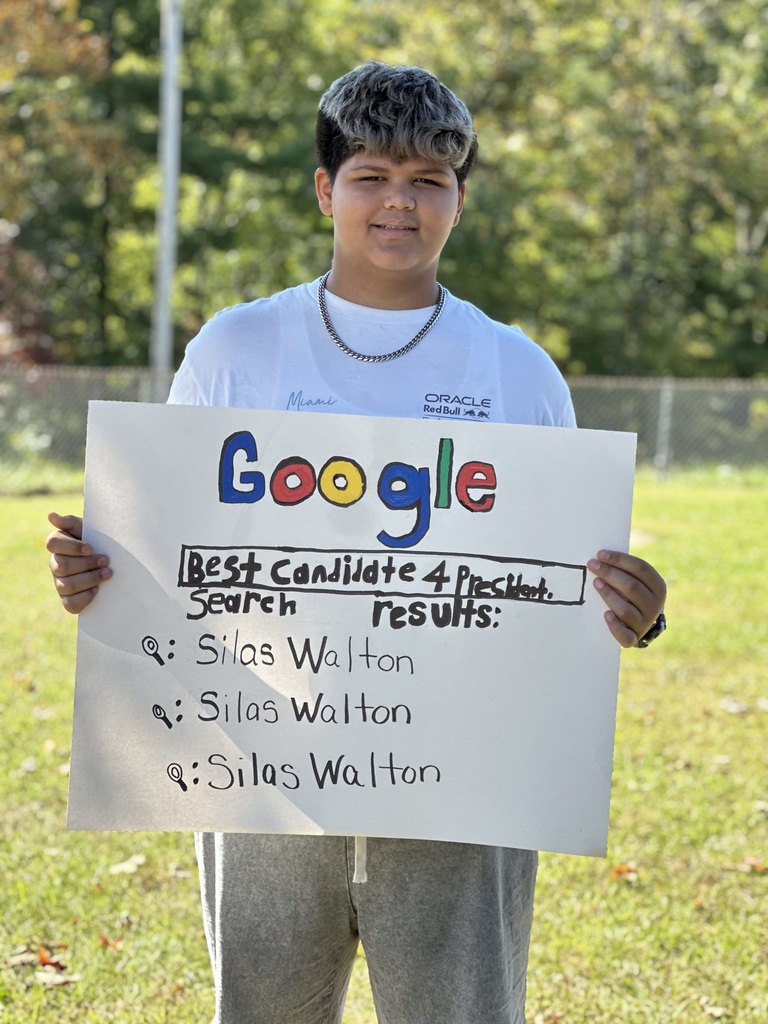 Boy holds a sign that says "Google" with "Best Candidate 4" and names listed. Trees and fence in background.