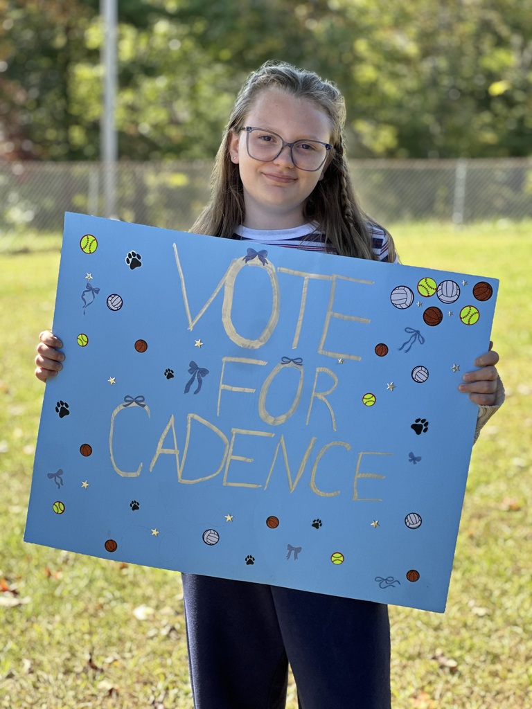 Girl wearing glasses and holding a blue sign that says, "Vote for Cadence" in front of a grassy area.