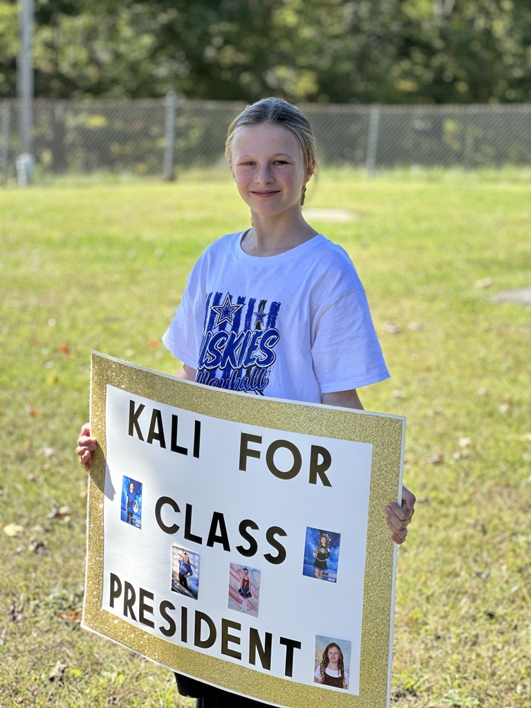 A young girl holding a campaign sign that says "Kali for Class President" stands in a grassy field.