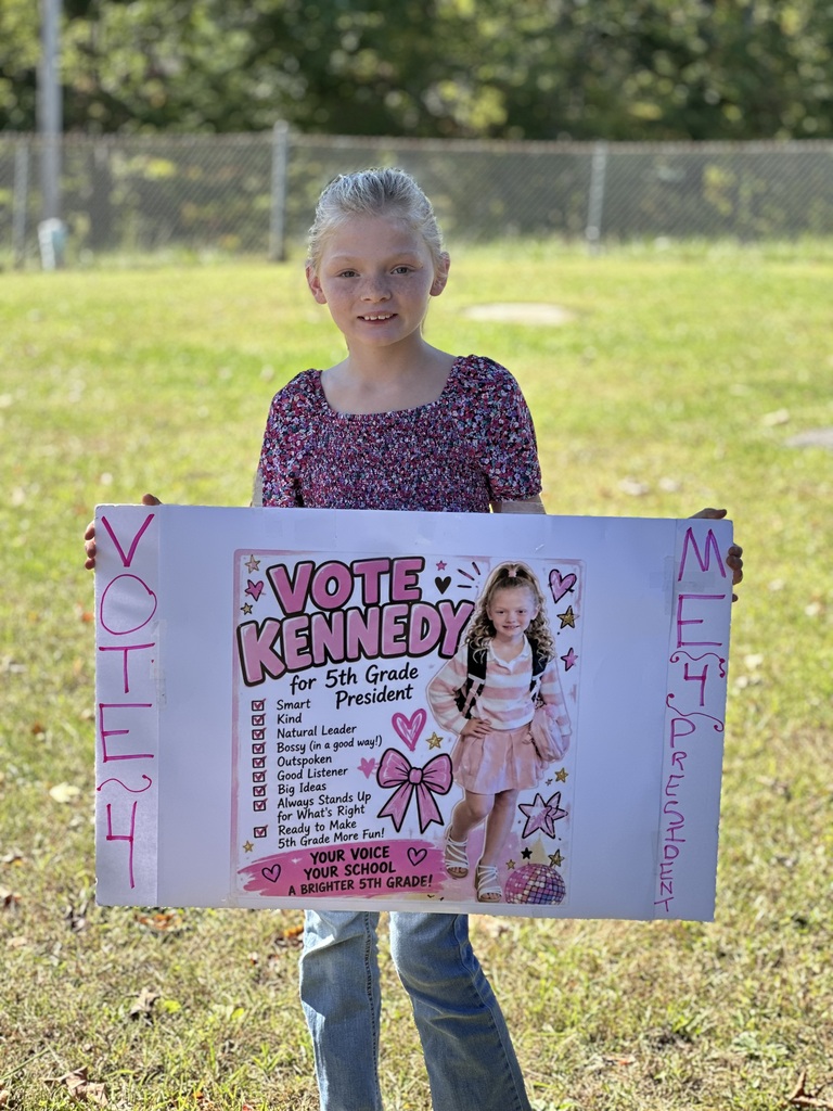A young girl holding a large poster with text and a doll, standing on a grassy field.