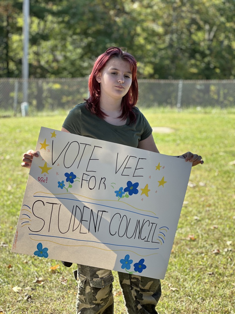 A woman with red hair holding a white sign that says "Vote Vee for Student Council" in blue and white letters.