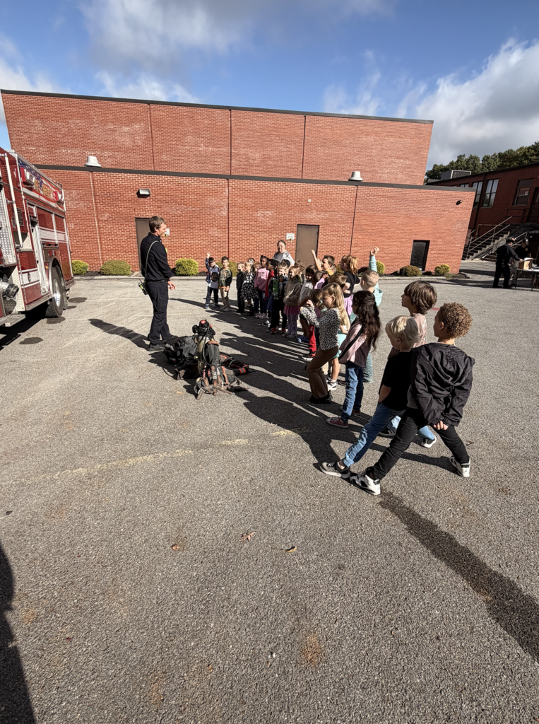 A group of children gather in a school parking lot. A fire engine is parked to the left.