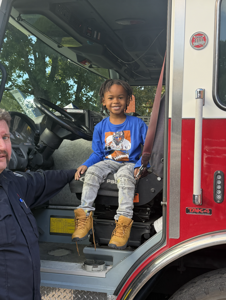 A man and a child in blue shirt sit in a fire truck. The child wears blue shirt, pants, and boots.