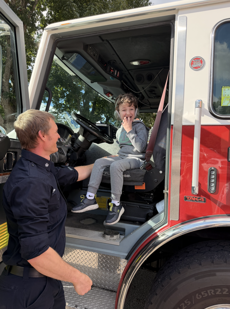 A firefighter helps a child sit in the cab of a fire truck. The child is wearing a blue hoodie.