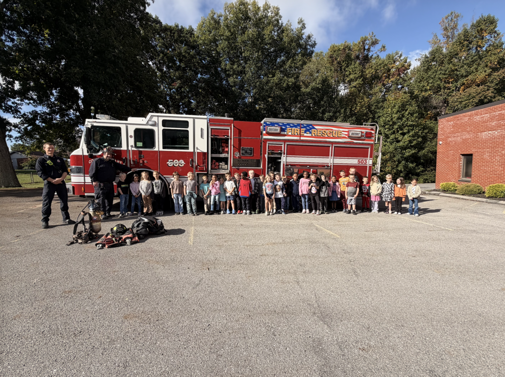 A group of people stands in front of a red firetruck, with two firefighters nearby. Trees and a building are in the background.
