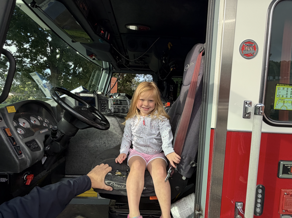 A child sits on a seat in a fire truck, with a hand holding the driver's door handle.