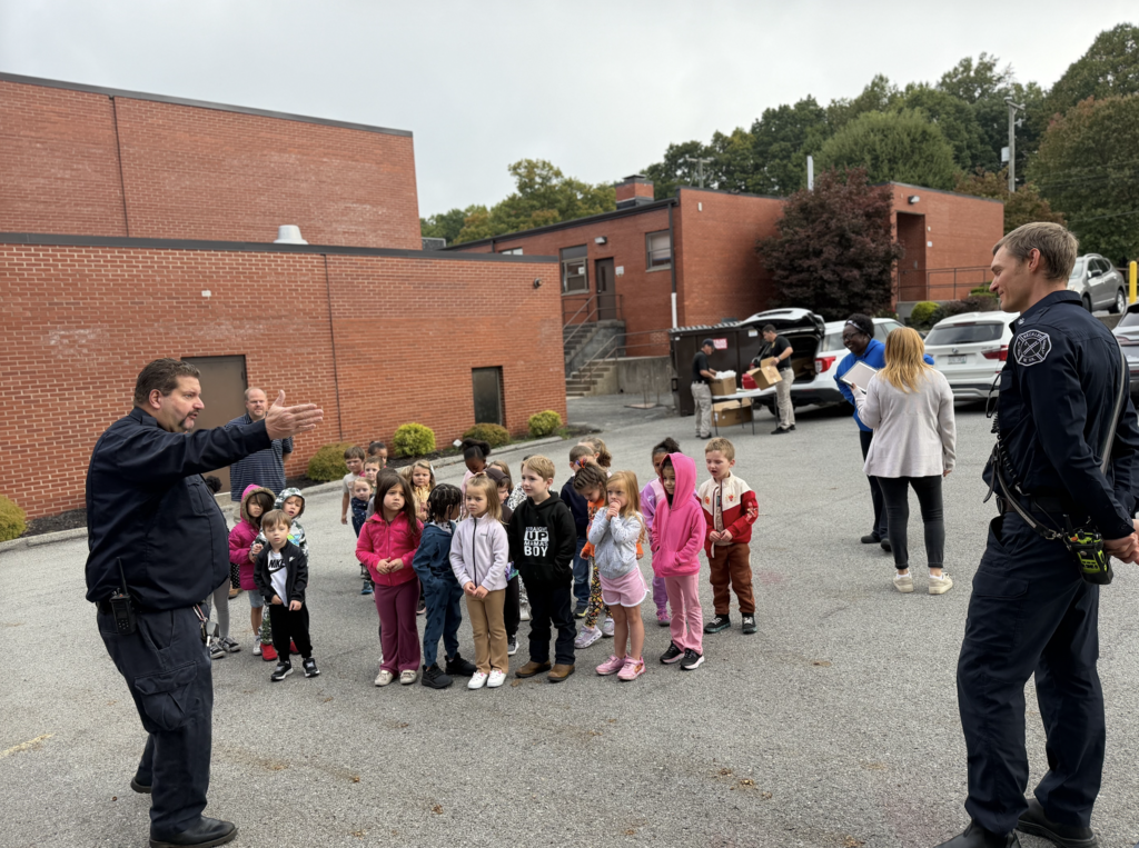 Police officers speak to a group of children in front of a building with trees and parked cars.