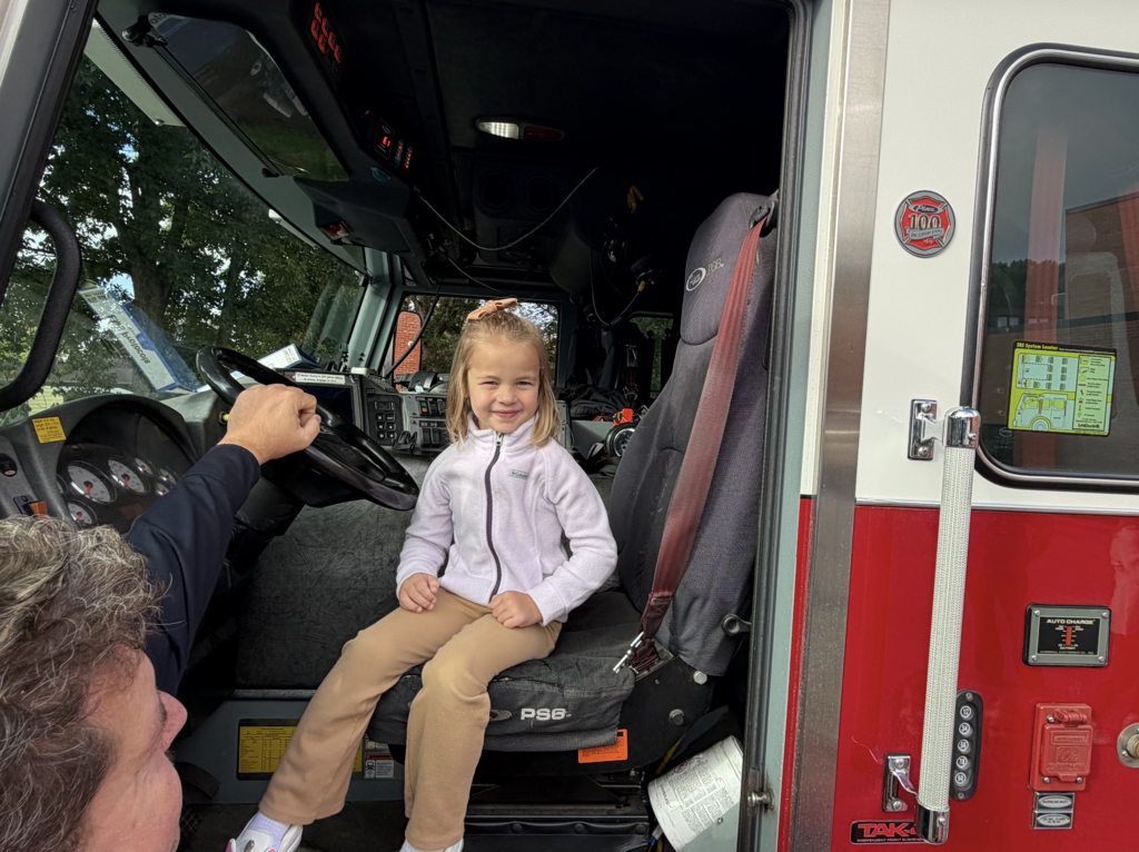 A young child sits in a firetruck driver's seat, with an adult in front holding the steering wheel.