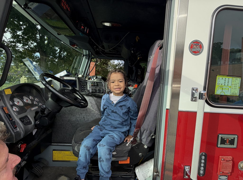 Child wearing blue pajamas sits in the driver's seat of an open fire truck.
