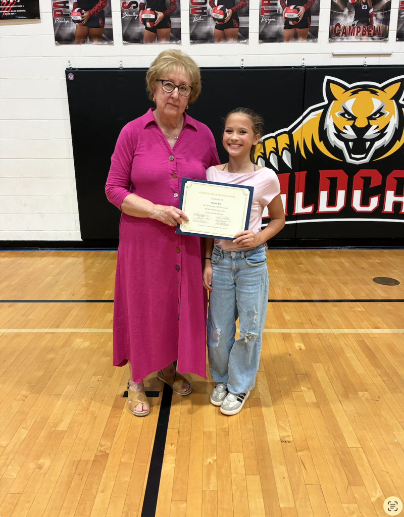 Woman in pink dress holds certificate next to young girl in jeans in gymnasium with tiger logo on wall.