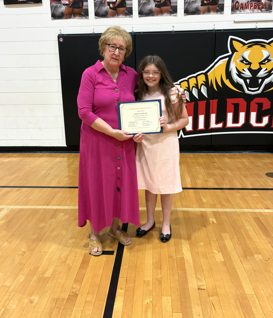A woman in a pink dress holds an award with a young girl in a pink dress. Behind them is a black wall with a tiger logo.