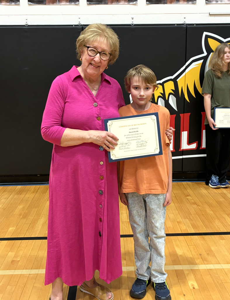 Woman in pink dress and boy in orange shirt holding framed certificate in a gymnasium.