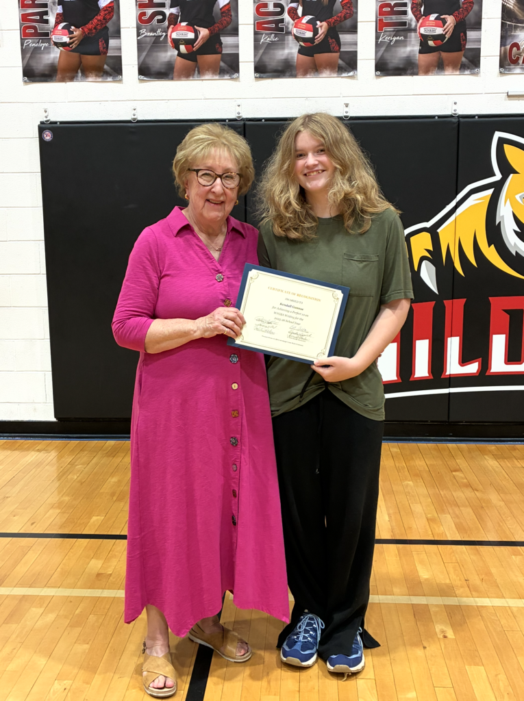 A woman in pink and a woman in green hold a certificate in a gym with a tiger logo.