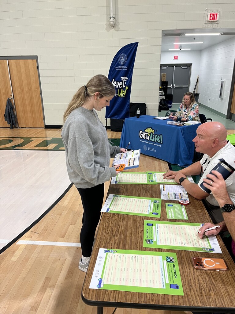A woman stands by a table with papers while a man sits nearby in a gymnasium.