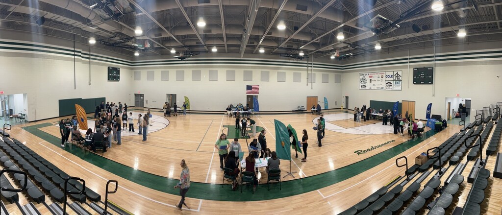 A basketball court with a crowd of people, green banners, and a flag. Bright lights are on the ceiling.