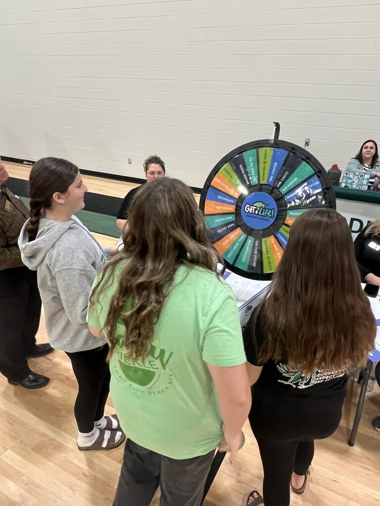 Women in a gymnasium engage with a colorful wheel game, with two in green and black shirts.