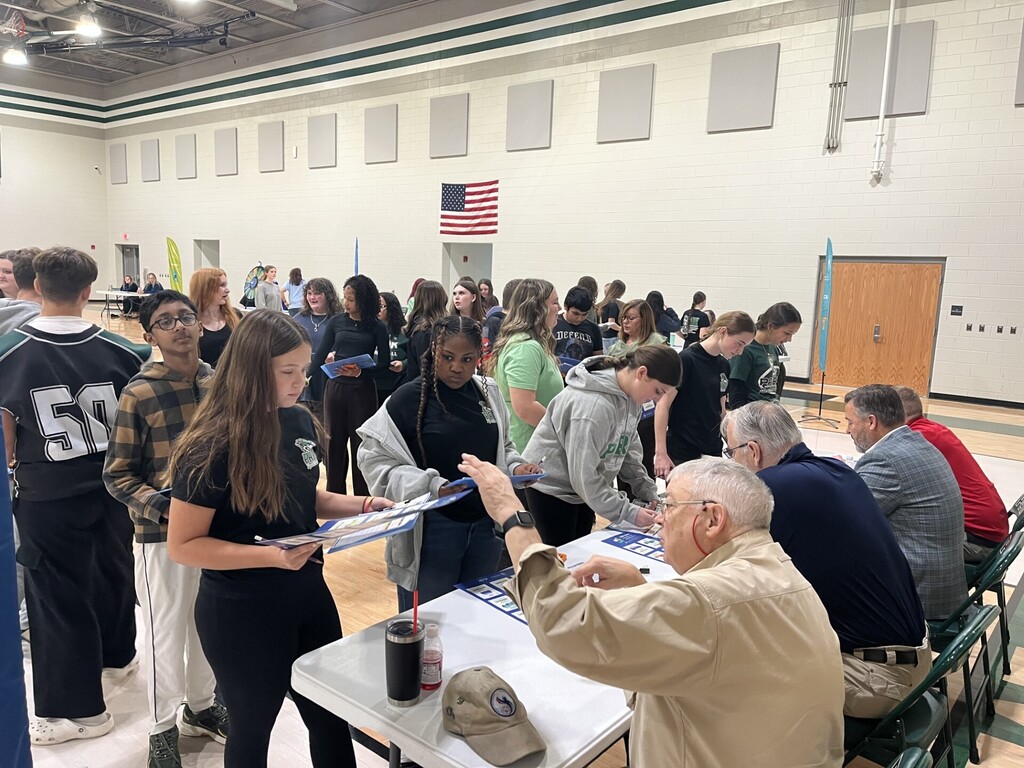 A gymnasium with tables and people seated or standing. A man hands a document to a woman.