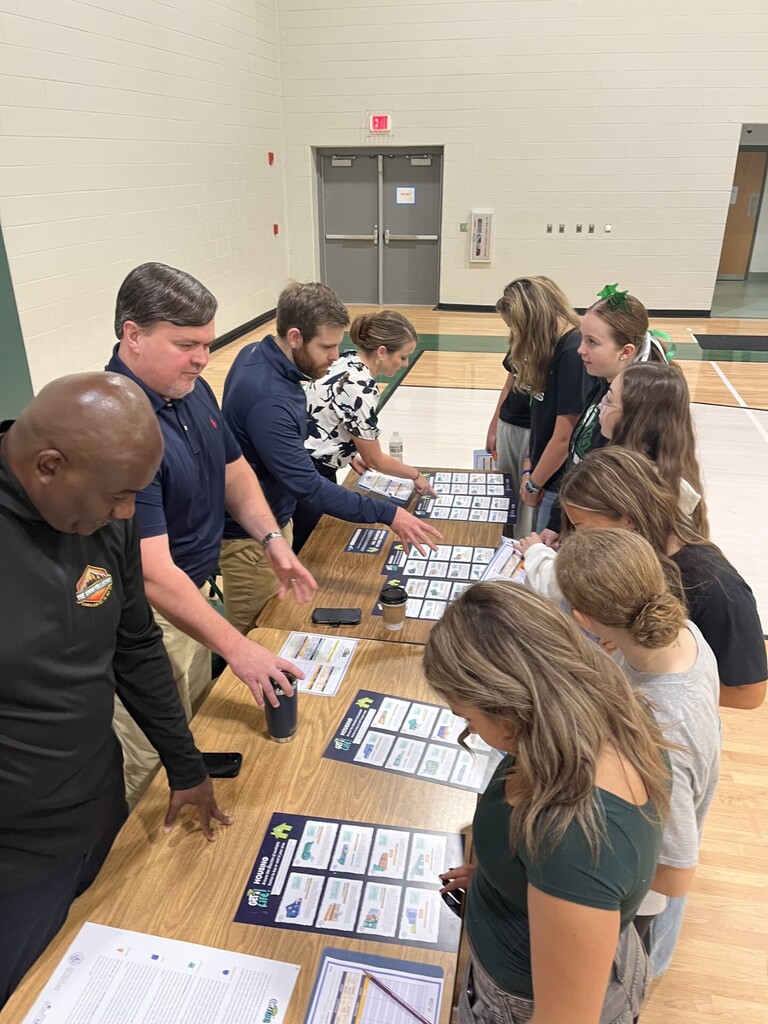 People gather around a long table with various cards, pamphlets, and a cup. Some are reading while others look at the cards.