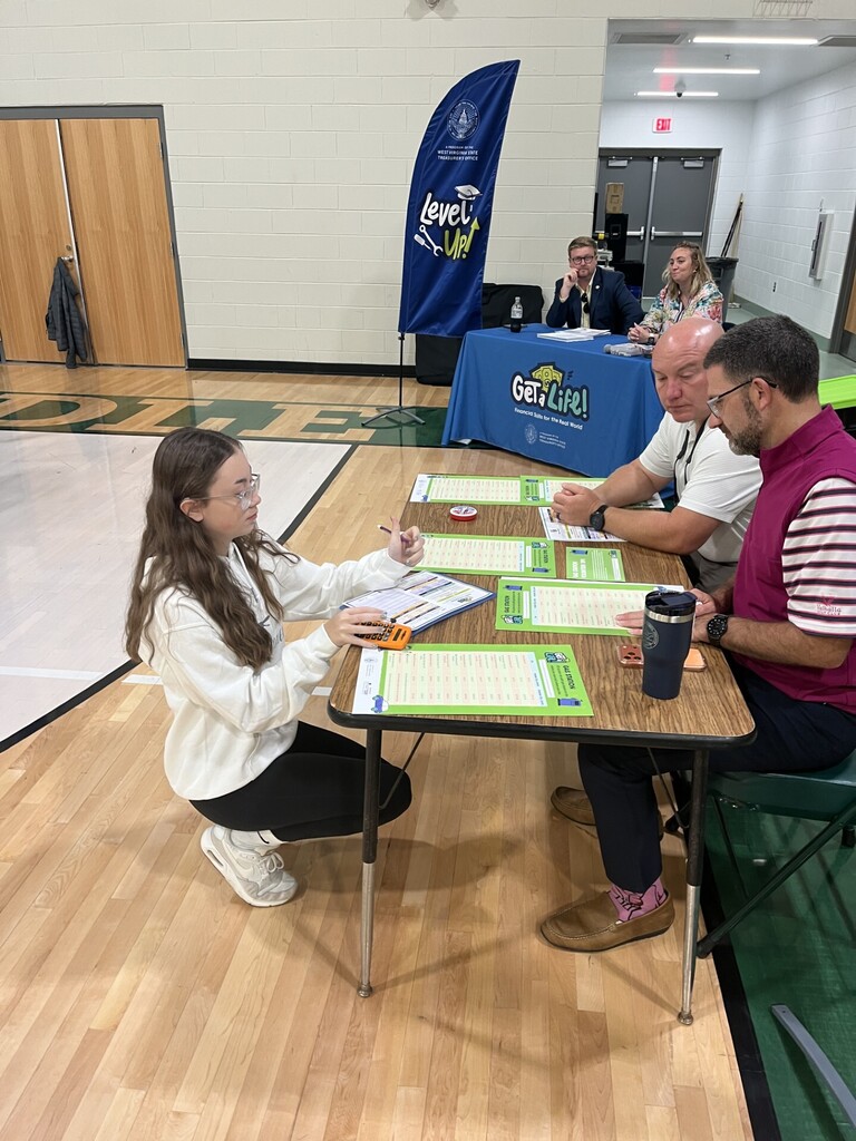 A girl kneels at a table with three adults. They are writing on paper with pens.