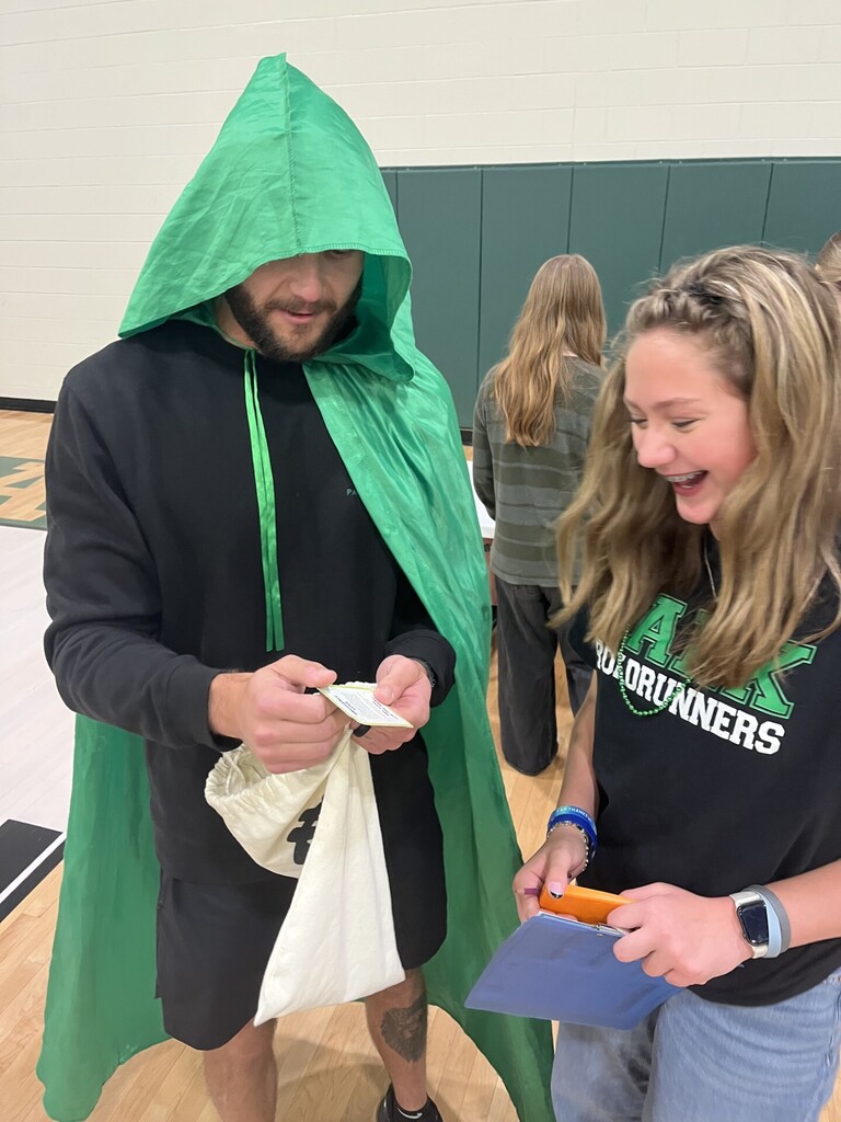 Person in a green cloak and a woman in a black shirt reading a note. Background: a school gym.